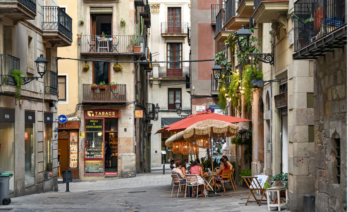 Un carrer estret de Ciutat Vella amb edificis antics amb balcons, una petita terrassa amb gent asseguda sota para-sols vermells i una botiga de tabacs al fons (font: Google)